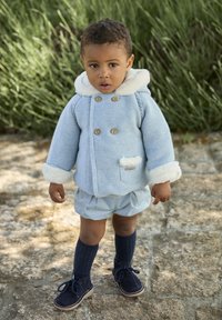 Abrigo de punto azul claro con ribete de piel blanca, botones de madera y bolsillo parche; combinado con pantalones cortos azul claro y calcetines y zapatos azul oscuro.