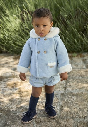 Abrigo de punto azul claro con ribete de piel blanca, botones de madera y bolsillo parche; combinado con pantalones cortos azul claro y calcetines y zapatos azul oscuro.