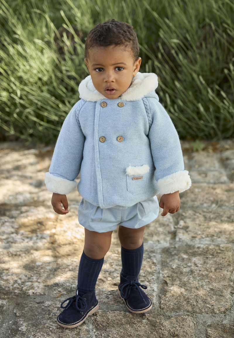 Abrigo de punto azul claro con ribete de piel blanca, botones de madera y bolsillo parche; combinado con pantalones cortos azul claro y calcetines y zapatos azul oscuro.