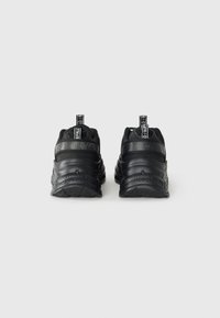 Baskets de sport noires avec un matériau texturé, forme arrondie et semelle prononcée. Présente une sangle noire marquée et une languette à l'arrière.