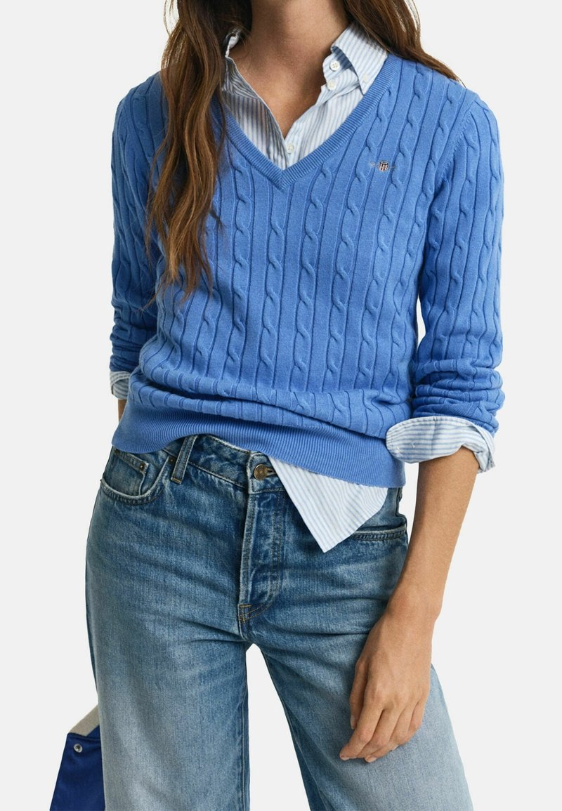 Femme portant un pull bleu en maille torsadée à col en V par-dessus une chemise à boutons rayée bleu et blanc, assorti d'un jean bleu délavé.