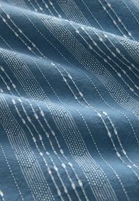 Tissu bleu avec des rayures verticales blanches texturées dans des motifs variés de points et de tirets, montrant des plis et des détails du matériau tissé doux.
