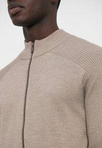 Béžový sveter na zips s rebrovanými detailmi na ramenách a golieri, s hladkou textúrou a priliehavým strihom. Viditeľný strieborný zips.