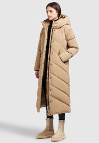 Manteau rembourré long beige avec un col haut, des boutons pression, un motif matelass�é diagonal et des poches latérales, associé à des bottes en caoutchouc beige.