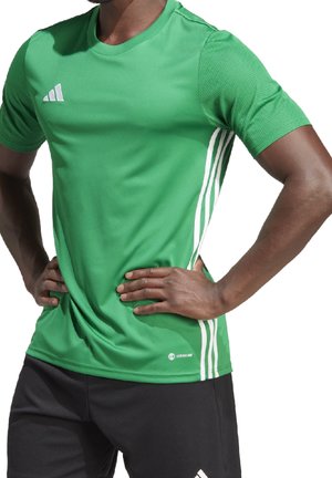 Mężczyzna w zielonej sportowej koszulce Adidas z białymi paskami i czarnych spodenkach, z rękami na biodrach, zwrócony w lewo.