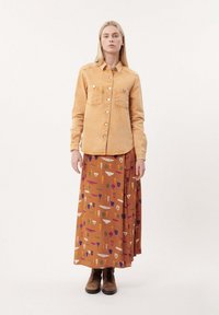 Beige denim shirt met knoopsluiting en dubbele borstzakken, gecombineerd met een lange oranjekleurige rok met kleurrijke abstracte patronen en bruine laarzen.