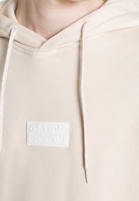 Beige värinen huppari pehmeästä kankaasta, jossa kengurutasku, valkoinen kohokuvioitu logo ja kontrastiväriset kiristysnauhat.