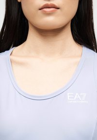 Κοντινό πλάνο στο κάτω μέρος του προσώπου και το λαιμό μιας γυναίκας που φοράει αμάνικο τοπ EA7 Emporio Armani σε ανοιχτό μοβ χρώμα, με ορατά τα κόκκαλα του λαιμού και ίσια σκουρόχρωμα μαλλιά.