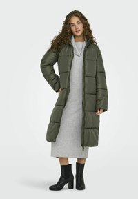 Dikke groene lange jas met een ritssluiting, gedragen over een lichtgrijze trui jurk. Zwarte enkellaarsjes maken de outfit compleet.