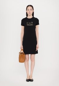 Μαύρο φόρεμα κοντό μανίκι με λογότυπο 'EA7 Emporio Armani' σε χρυσό, σε συνδυασμό με μια πλεκτή τσάντα σε καφέ απόχρωση. Διαθέτει ίσια γραμμή και στρογγυλή λαιμόκοψη.