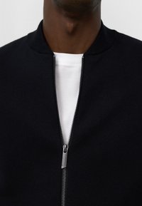 Man i en delvis uppdragen svart zip-up-tröja över en vit t-shirt, synlig från hals till bröst.