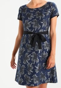 Granatowa sukienka z krótkim rękawem, w kwiatowy wzór, z wcięciem w talii i czarnym paskiem. Sylwetka w kształcie litery A, z miękką teksturą i subtelnym połyskiem.