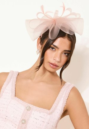 Ung kvinde iført lyserød, tekstureret, ærmeløs kjole med rhinstenknapper og stort, gennemsigtigt lyserødt sløjfe-hovedbånd, neutral baggrund.