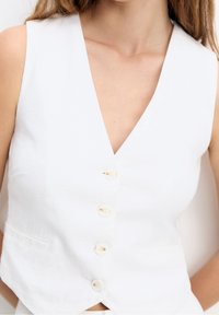 Gilet sans manches blanc avec un décolleté en V prononcé, comportant cinq boutons recouverts de tissu, une finition texturée et deux poches latérales.