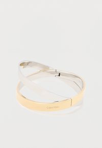 Verschlungene, polierte Metallarmreifen in Silber- und Goldtönen, einer eingraviert mit dem "Calvin Klein"-Logo, auf einem weißen Hintergrund.