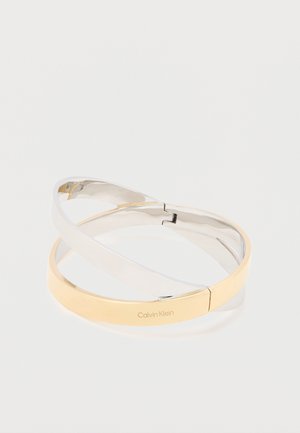 In elkaar grijpen gepolijste metalen armbanden in zilver- en goudtinten, waarvan er één is gegraveerd met het "Calvin Klein"-logo, op een witte achtergrond.