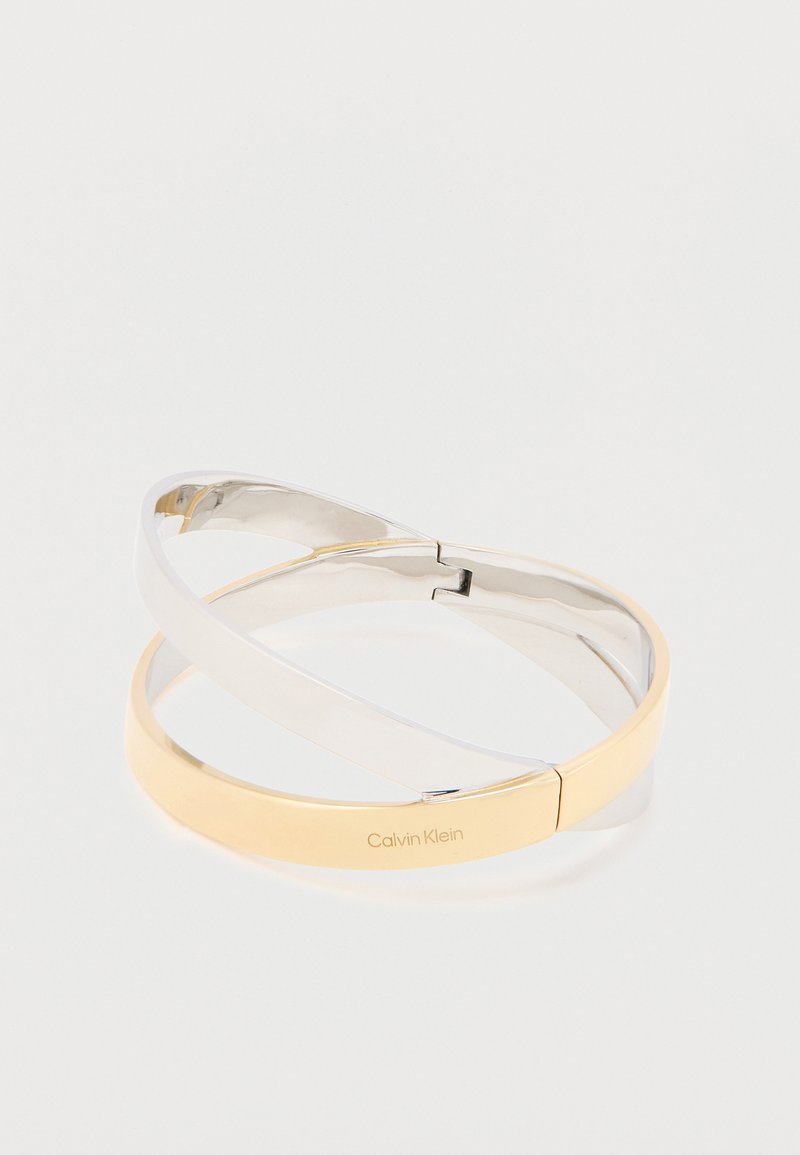 Verschlungene, polierte Metallarmreifen in Silber- und Goldtönen, einer eingraviert mit dem "Calvin Klein"-Logo, auf einem weißen Hintergrund.