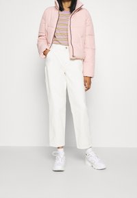 Giubbotto trapuntato rosa chiaro con colletto alto, indossato sopra una maglietta a righe viola. Abbinato a pantaloni bianchi a gamba larga e sneakers bianche.