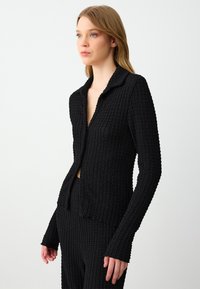 Zwarte gestructureerde gebreide cardigan met een kraag, lange mouwen en een open voorkant. Heeft een getailleerde pasvorm en ribbel details.