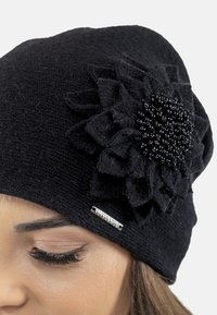 Bonnet tricot noir avec un accent de fleur texturé, orné de perles et d'une petite étiquette en métal sur le côté. Tissu doux et design ajusté.