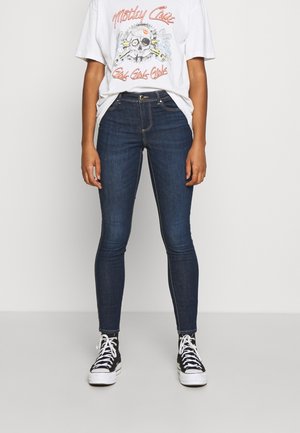 Persoon die donkerblauwe skinny jeans draagt, zwarte hoge sneakers met witte veters, en een wit T-shirt met een schedel- en tekstontwerp.