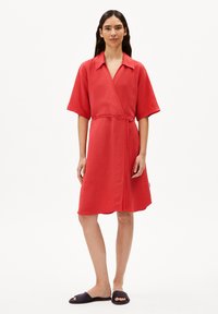 Rotes Wickelkleid aus leichtem Stoff, mit kurzen Ärmeln und gebunden in der Taille. Verfügt über einen Kragen und endet knapp über dem Knie. Dunkle Riemensandalen.