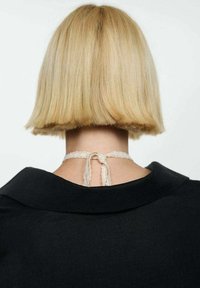 Tăietura de păr bob scurt, blond, văzută din spate, cu o brăță din dantelă delicată legată la ceafă, persoană îmbrăcată într-o haină neagră cu guler larg.