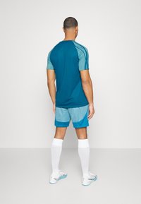 Man i teal sporttröja och ljusblå shorts står vänd bort mot en enkel vit bakgrund.