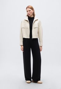 Κρεμ cropped μπουφάν με δύο μπροστινές τσέπες, κουμπιά και δομημένο γιακά, συνδυασμένο με μαύρο παντελόνι φαρδιάς ποδής και μπεζ παπούτσια.