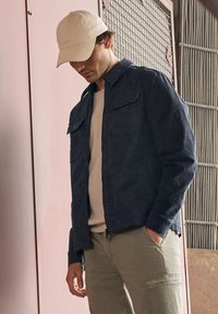 Giacca blu navy con texture e tasche con bottoni, abbinata a una camicia beige chiaro e pantaloni cargo oliva, completata da un berretto beige chiaro.