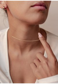 Un choker en or repose légèrement sur le cou, fin et minimal dans son design. Un doigt touche doucement le collier, mettant en valeur sa forme délicate.