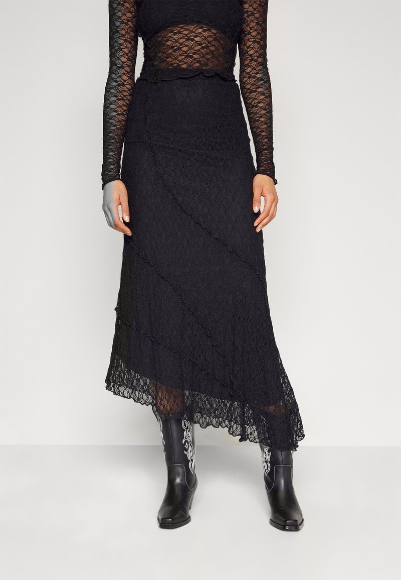 Femme portant un haut en dentelle noire à manches longues et une jupe en dentelle noire assortie, avec des bottes de cowboy noires brodées en blanc.