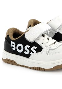 Sneaker bianchi e nere con una suola marrone; presentano una strap in velcro, pannelli laterali texturizzati e un logo "BOSS" prominente.