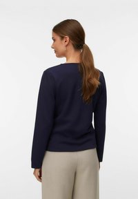 Mornarsko modra majica z dolgimi rokavi in okroglo ovratnikom, v kombinaciji s svetlobezžimi hlačami, ki imajo gladko teksturo tkanine in oprijet kroj.