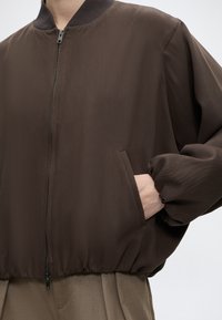Personne portant une veste marron à fermeture éclair avec col côtelé et poignets élastiques, main dans une poche avant, associée à un pantalon marron.