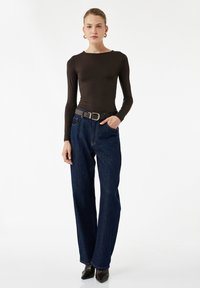 Een bruine longsleeve top gecombineerd met losse donkerblauwe denim jeans en een zwarte riem met zilveren gesp, en puntige zwarte hakken.