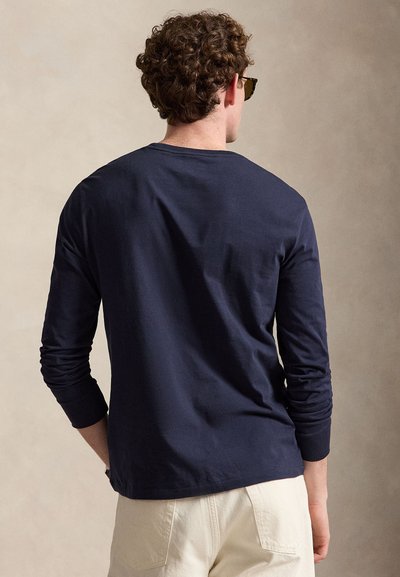 Göndör hajú férfi napszemüvegben, tengerészkék hosszú ujjú ingben és bézs nadrágban, egyszínű bézs háttér előtt áll.