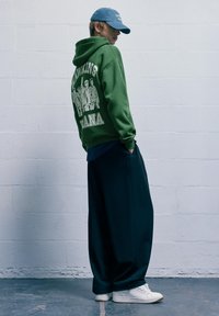 Zelená mikina s bielym grafickým dizajnom na chrbte, doplnená tmavými voľnými nohavicami a bielymi teniskami. Modrá čiapka dotvára outfit.