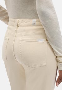 Beżowe dżinsy z denimu z tylnymi kieszeniami, dopasowaną talią i widocznym przeszyciem. Na prawej kieszeni znajduje się mała metka z logo marki.