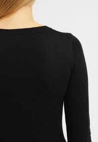 Suéter negro de punto con escote redondo, diseño ceñido y puños acanalados en las mangas. Textura suave, con un detalle mínimo en la parte posterior.