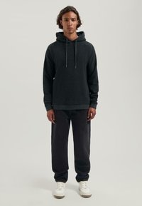 Giovane uomo in piedi rivolto frontalmente, che indossa un felpa grigia scura, jeans neri e sneakers bianche, su uno sfondo chiaro e semplice.