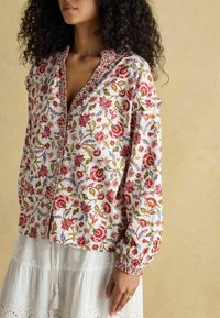 Kwiatowa bluzka z białej tkaniny z czerwonymi i zielonymi wzorami, zapinana na guziki z przodu, z bufkami. Noszona na białej spódnicy z falbanami.