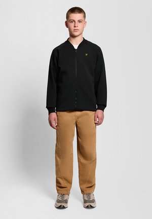 Jeune homme debout face à l'avant, portant une veste noire à fermeture éclair, un pantalon beige et des baskets beige sur un fond gris uni.