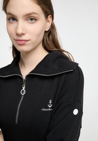 Zwarte rits hoodie met een grote kraag, voorzien van een zilveren rits, geborduurd ankerlogo en een witte knop op de mouw.