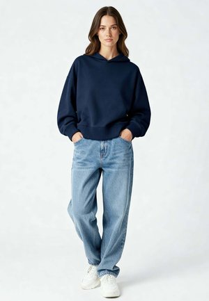 Jonge vrouw staand met handen in zakken, draagt een marineblauwe hoodie, losse lichtblauwe spijkerbroek en witte sneakers tegen een effen achtergrond.