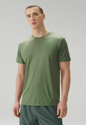 Junger Mann mit kurzem Haar, der ein eng anliegendes, gemustertes olivgrünes T-Shirt und dunkelgrüne Shorts trägt und vor einem schlichten, hellen Hintergrund steht.