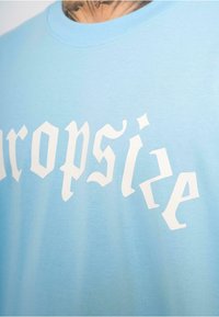 Lyseblå bomulds-t-shirt med fed, stiliseret hvid tekst. Udhullet er rundt, og stoffet ser blødt ud med en glat tekstur.