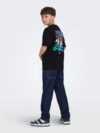 Pojke som bär svart t-shirt med färgglatt blommönster på ryggen, mörkblå jeans och blåvita sneakers, stående i profil mot vit bakgrund.