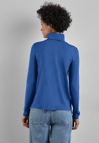 Blaues Langarmoberteil mit Stehkragen. Weiche Stoffstruktur, taillierte Passform. Kombiniert mit hoch taillierten hellen Jeans.