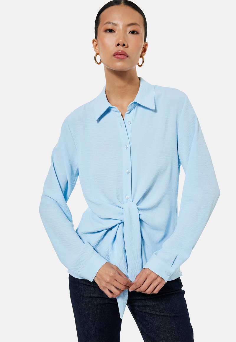 Mujer con blusa azul claro abotonada y anudada en la cintura, pantalones oscuros y grandes pendientes de aro, de pie con las manos entrelazadas.